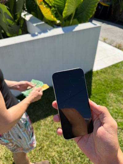 A used iPhone with a scratched screen held up on a back lawn while someone counts out notes beside it