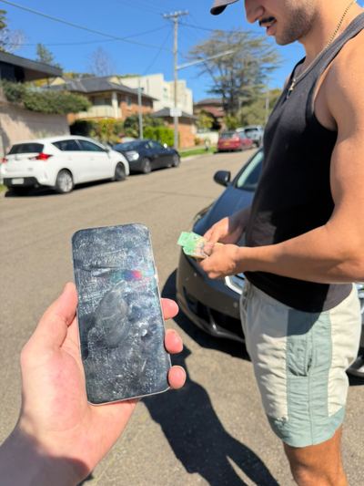A man counting out notes on a suburban street while a used iPhone with a badly scratched screen is held up to the camera