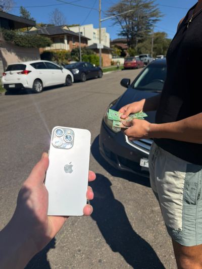 A used iPhone held up in one hand in the middle of a suburban street, with notes being counted beside a car behind it