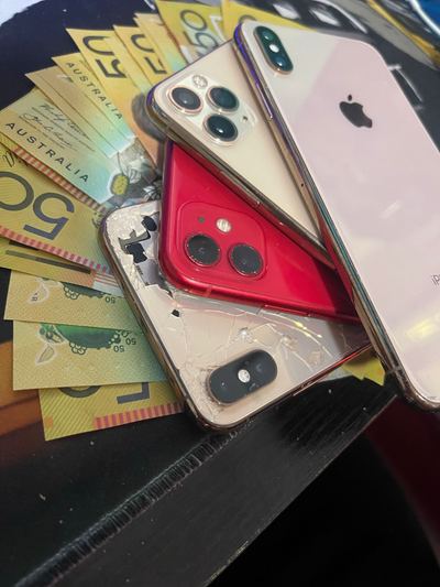 Four used iPhones stacked on a workbench mat over a fan of fifty dollar notes, the one underneath with its back glass in pieces